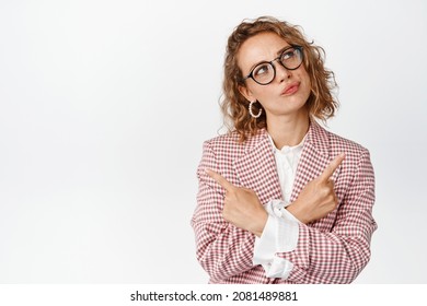 Thoughtful business woman in glasses and stylish suit, looking intrigued and pointing sideways, making decision, thinking, picking from variants, white background