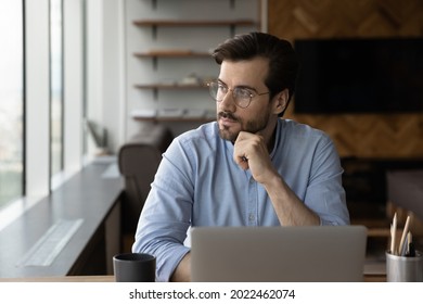 Thoughtful businessman in glasses looking at window from workplace with laptop, thinking over challenges, work issues, problem solving, making decision, feeling doubts, pondering on future vision - Powered by Shutterstock