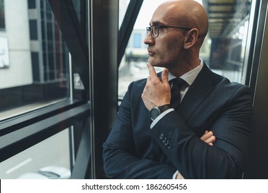 Thoughtful Businessman In Suit In Eye Glasses Looking Away In The Window Or Glass Elevator With A Busy Street View. Focused Business Man Face Looking Outside Of The Window. 