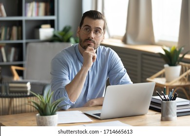 Thoughtful businessman wearing glasses touching chin, pondering ideas or strategy, sitting at wooden work desk with laptop, freelancer working on online project, student preparing for exam at home - Shutterstock ID 1714665613