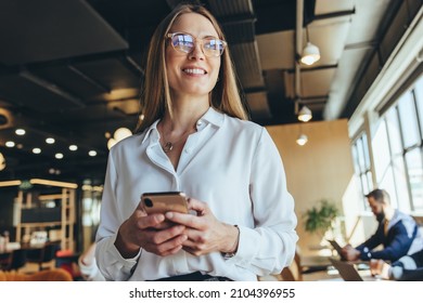 Thoughtful businesswoman holding a smartphone in a co-working space. Happy young businesswoman looking away with a smile while standing in a modern workplace. Businesswoman thinking of new ideas. - Shutterstock ID 2104396955