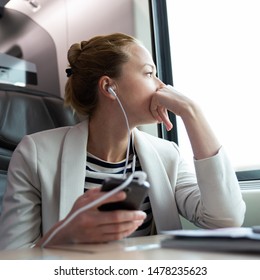 Thoughtful Businesswoman Looking Trough The Window, Listening To Podcast On Cellphone Using Headphone Set While Traveling By Train In Business Class Seat.