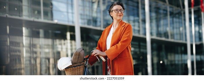 Thoughtful businesswoman pushing her bike during her morning commute in the city. Mature business woman looking away pensively while walking to work with a bicycle. - Powered by Shutterstock