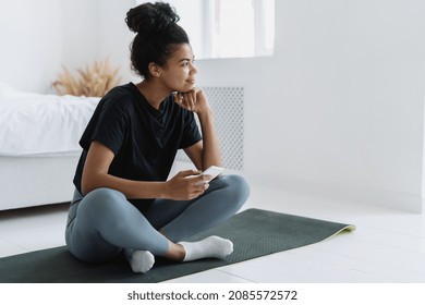 Thoughtful and calm african ethnicity woman holding smartphone in her hand, sitting on yoga mat and resting after sport training. Female enjoying morning at home - Powered by Shutterstock