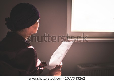 Thoughtful cancer ill woman reading her medical reports and looking through the window. 