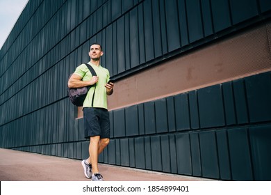 Thoughtful Caucasian Male Bodybuilder In Active Sportswear Walking At Urban Setting Going To Cardio Training, Muscular Sportsman With Backpack And Smartphone Technology Thinking About Physical Workout