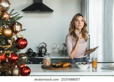 Thoughtful Caucasian woman with blond hair casual outfit having healthy breakfast writing making notes to do list standing in the cozy kitchen with Christmas tree in the foreground front. - Powered by Shutterstock