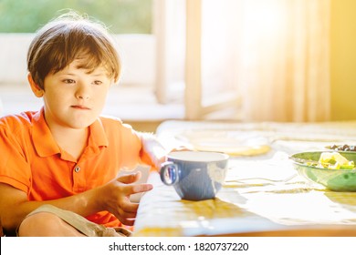 Thoughtful Child Boy Wearing Orange T-shirt With Down Syndrome Sitting At Table At Home. Sun Glare Effect. Special Needs Children Concept.