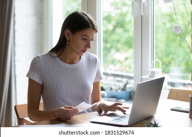 Thoughtful concentrated young woman holding paper bills in hands, entering payment information in computer application, paying utility expenditures online using laptop software alone at home.
