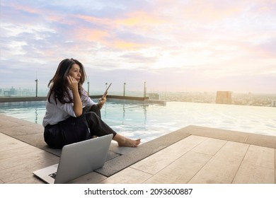 Thoughtful Concerned Asian Woman Working On Laptop And Using Tablet Looking Away Thinking Solving Problem In Evening At Pool On Skyscraper, Serious Woman Search For Inspiration. Copy Space 