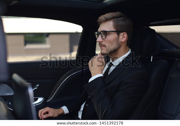 Thoughtful confident businessman keeping hand
on glasses while sitting in the luxe
car.