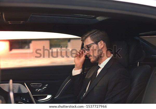 Thoughtful confident businessman keeping hand\
on glasses while sitting in the luxe\
car.