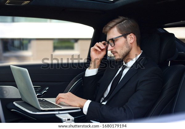 Thoughtful confident\
businessman keeping hand on glasses while sitting in the luxe car\
and using his\
laptop.