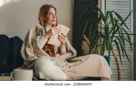 Thoughtful and cozy red-haired woman sitting with her legs crossed and taking notes on notebook. Relaxation lifestyle concept - Shutterstock ID 2238403693