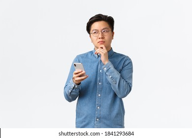 Thoughtful creative asian man in glasses thinking while making post on social media, looking away, pondering or making decision, holding smartphone, choosing something in internet