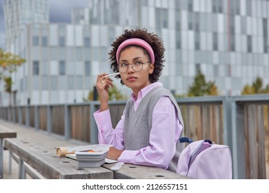 Thoughtful Curly Haired Clever Female Student Writes Down Notes In Notebook Creats Essay Poses Outdoors Against Blurred Background Focused Pensively Into Distance Surrounded By Lunchbox And Rucksack