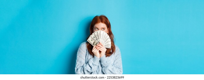 Thoughtful cute girl with red hair dreaming about shopping, holding dollars and looking at upper left corner logo, standing over blue background. - Shutterstock ID 2253823699