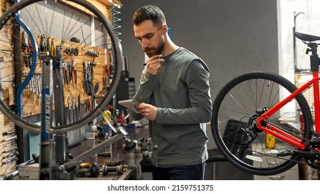 Thoughtful Cycling Repairman Reading Or Writing Notes In Notebook During Working In Workshop. Young Caucasian Bearded Man. Bike Service, Repair And Upgrade. Garage Interior With Tools And Equipment