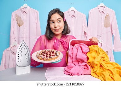 Thoughtful Dark Haired Woman Looks Aside Thinks About Something Takes Break After Doing Domestic Chores Stands Near Ironing Board With Pile Of Laundry And Electric Iron Holds Plate Of Tasty Pie