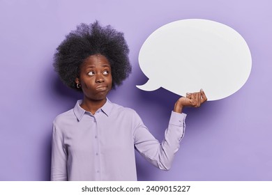 Thoughtful dark skinned beautiful young woman with curly bushy hair wears shirt holds blank speech bubble for your text advertisement area isolated over purple background. Your message canvas - Powered by Shutterstock