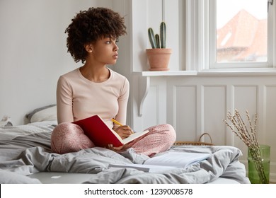 Thoughtful Dark Skinned Young Woman Writes At Textbook, Sits In Lotus Pose, Wears Nightwear, Sits In Cozy Bedroom Alone, Thinks About Creative Ideas For Writing Essay, Focused Aside In Window