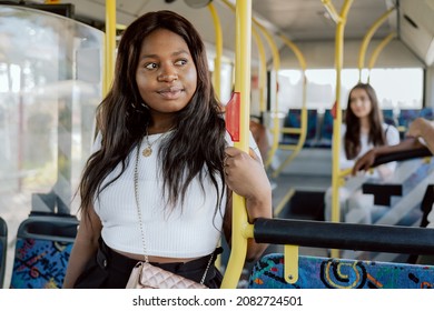 Thoughtful dark-skinned woman is riding bus, looking out window standing by seats, holding onto railing, looking out for the stop, analyzing schedule, the way to work by public transportation