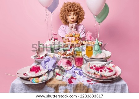 Thoughtful displeased birthday girl poses near festive table variety of desserts feels lonely on party poses against pink wall with inflated balloons near. Unhealthy sweet food for special occasion