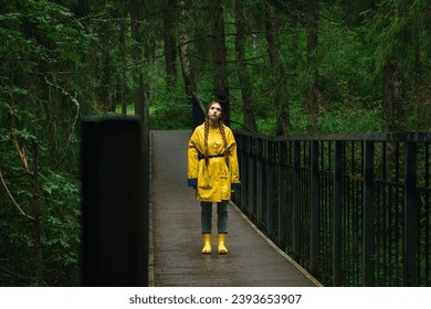 Thoughtful and dreaming woman in a yellow raincoat and rubber boots stands on a bridge in a green wet park during the rain. The concept of travel and tourism. - Powered by Shutterstock