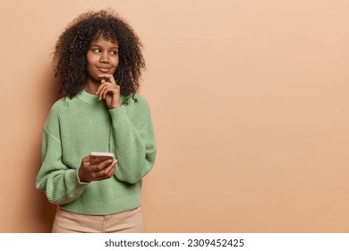 Thoughtful dreamy Afro woman with curly hair keeps hand on chin considers something uses mobile phone dressed casual green knitted jumper and trousers isolated over beige background copy space - Shutterstock ID 2309452425