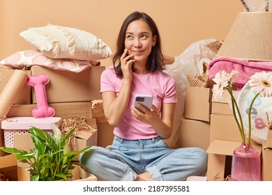 Thoughtful dreamy Asian woman uses modern smartphone poses in messy room around boxes full of personal belongings moves in new apartment or searches room to rent in internet. Relocation concept - Shutterstock ID 2187395581