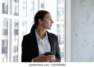 Thoughtful Dreamy Lost In Thoughts Young Businesswoman, Or Company Manager In Formal Suit Holding Smartphone Looking Out Window At Big City View. Business Vision, Future Opportunities, Tech Concept