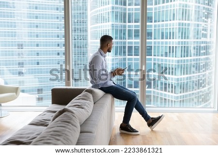 Thoughtful dreamy millennial Black man using smartphone in office lobby, hotel room, at home, looking at city building view in window away, thinking of online chat message against glass background