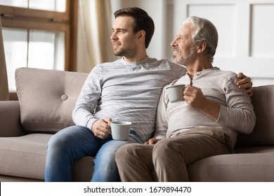 Thoughtful Elderly Father And Adult Son Sit Relax On Couch In Living Room Drink Tea Look In Distance Dreaming, Pensive Dreamy Mature Adult And Grown-up Child Rest On Sofa At Home Visualizing Together