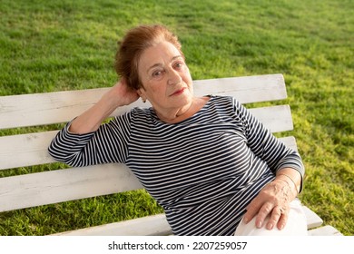 Thoughtful Elderly Woman, Pensioner Relaxed. Portrait Of Positive Granny On A Bench In The Park. Sunny Day. Concept Of Old Age, Health Insurance. Life Style. Outdoor. Caucasian Lady 80+. Green Grass.