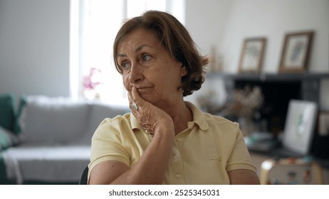 Thoughtful elderly woman resting her chin on her hand while sitting indoors, looking away with a pensive expression, wearing a yellow polo shirt, home background visible, natural light - Powered by Shutterstock