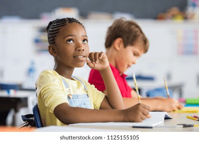 Thoughtful Elementary School Girl Sitting At Desk And Looking Up. Pretty Schoolgirl Looking Up During Exam With Classmate In Background. Black Scholar Thinking About The Solution Of The Exam.