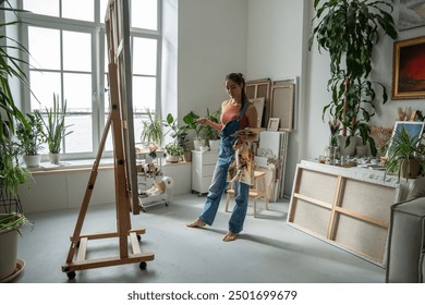 Thoughtful female artist illustrator stands and mixes paints on palette, contemplating idea of new painting on easel in bright art studio or drawing school. Beginning of new work of art in art school - Powered by Shutterstock