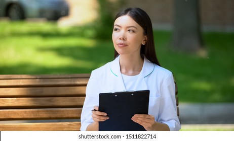 Thoughtful Female Doctor Working Outside, Sitting On Bench In Hospital Park