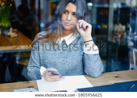 Thoughtful female novelist in grey sweater adjusting glasses and looking out window while browsing internet and writing book from cozy time cafe