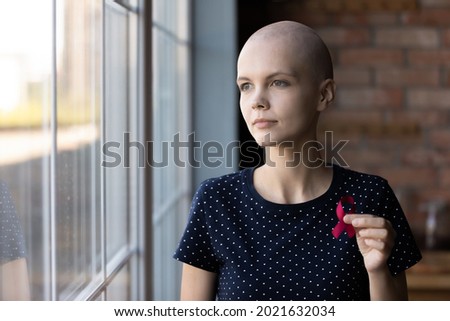 Thoughtful female oncology patient holding red ribbon, showing symbol of fight against breast cancer, expressing support and solidarity to ill women, promoting early medical checkup and diagnosis