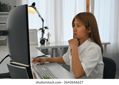 A Thoughtful female programmer concentrates on the computer while coding in a modern office setup.