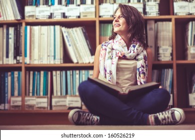 Thoughtful female student sitting against bookshelf with a book on the library floor - Powered by Shutterstock