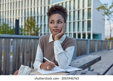 Thoughtful female student writes notes in notebook holds pen has pensive expression puts down her thoughts in personal diary during leisure time wears neat clothes makes schedule records checklist