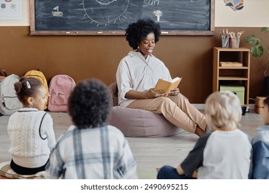 Thoughtful female teacher of Black ethnicity in bean bag chair reading book aloud to primary school students sitting around on classroom floor - Powered by Shutterstock