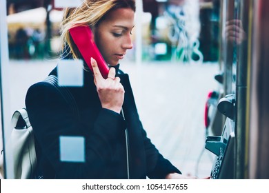 Thoughtful Female Tourist Making Call In Payphone On Street Standing In Transparent Booth, Young Woman Using Public Telephone Operated With Coins During Travel For Call With Low Prices Abroad