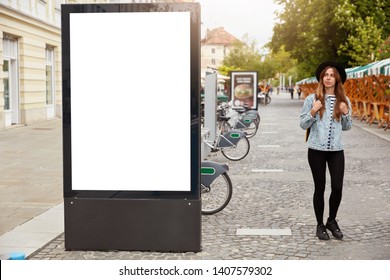 Thoughtful female tourist strolls on footway near lightbox with mock up blank space for your advertising content or commercial information. Street style concept. Focus on billboard at sidewalk