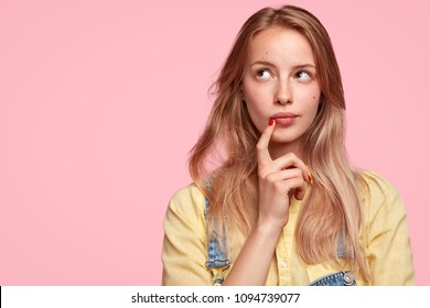 Thoughtful female youngster keeps fore finger near lips, being deep in thoughts, thinks about coming holidays, isolated on pink background with blank copy space for your advertisement or text - Powered by Shutterstock