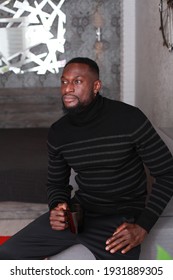 Thoughtful Fit Muscular Black Man In Sweater With Turtle Neck Sitting On Coach. Domestic Photo At Home. People Lifestyle Concept. Handsome Black Man's Portrait. Male Self-Confidence Concept