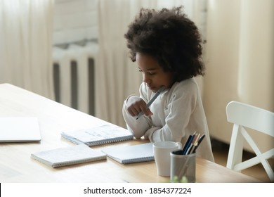 Thoughtful Focused African American Little Girl Studying At Home, Sitting At Table, Holding Pen, Pensive Dreamy Girl Child Pondering Difficult School Tasks, Doing Homework, Homeschooling Concept