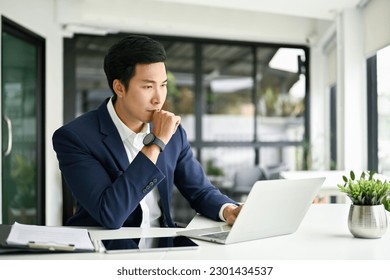 Thoughtful and focused millennial Asian businessman or male boss is pensively thinking about a plan to solve his project's problems, looking at his laptop screen, and working in his office. - Powered by Shutterstock